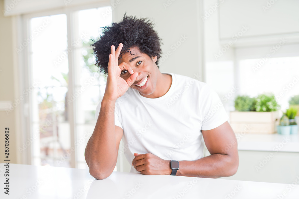 Young african american man wearing casual white t-shirt sitting at home doing ok gesture with hand smiling, eye looking through fingers with happy face. Young african american man wearing casual white t-shirt sitting at home doing ok gesture with hand smiling, eye looking through fingers with happy face.