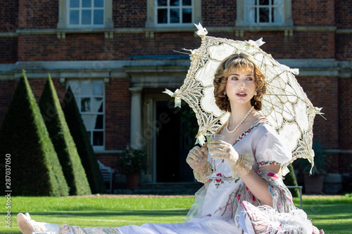Beautiful lady in regency clothing sitting on lawn in front of stately home holding parasol and drinking sparkling wine photo