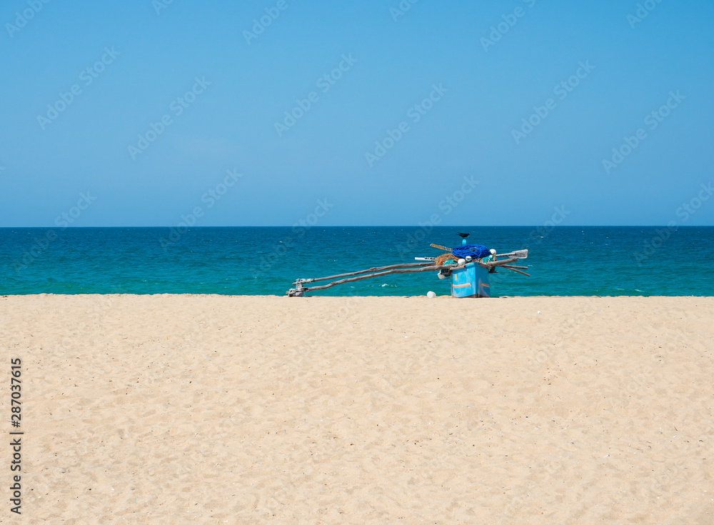 Traditional wooden fishing boat on empty beach against clear blue sky and sea, Kallady Beach, Batticaloa, Sri Lanka.