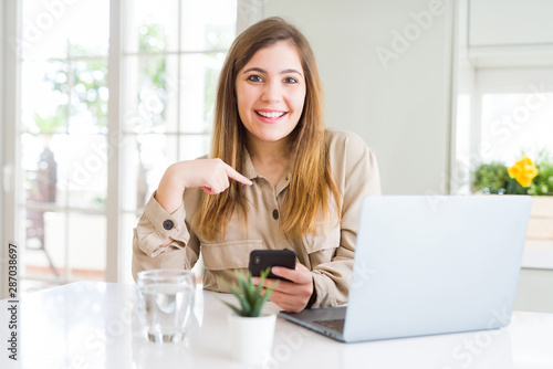 Beautiful young woman using smartphone and computer with surprise face pointing finger to himself