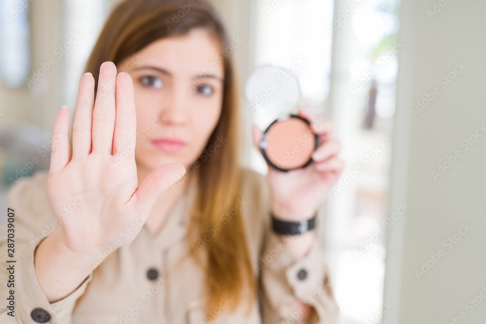 Beautiful young woman using make up cosmetics applying powder with open hand doing stop sign with serious and confident expression, defense gesture Beautiful young woman using make up cosmetics applying powder with open hand doing stop sign with serious and confident expression, defense gesture