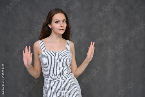 Portrait of a cute brunette girl, a young woman with beautiful curly hair in a gray dress on a gray background. Smiling, talking with emotions, showing hands to the sides.