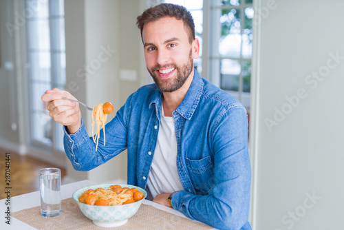 Handsome man eating pasta with meatballs and tomato sauce at home while smiling at the camera