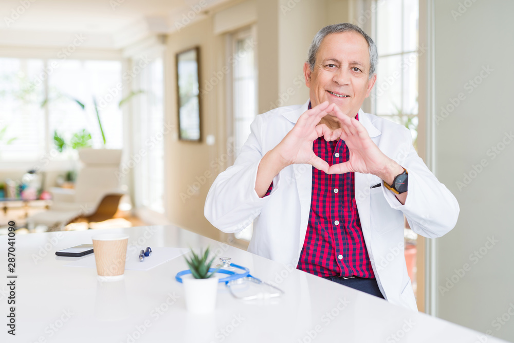 Handsome senior doctor man wearing medical coat at the clinic smiling in love showing heart symbol and shape with hands. Romantic concept. Handsome senior doctor man wearing medical coat at the clinic smiling in love showing heart symbol and shape with hands. Romantic concept.