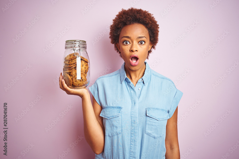 African american woman holding jar of chocolate chips cookies over pink isolated background scared in shock with a surprise face, afraid and excited with fear expression African american woman holding jar of chocolate chips cookies over pink isolated background scared in shock with a surprise face, afraid and excited with fear expression