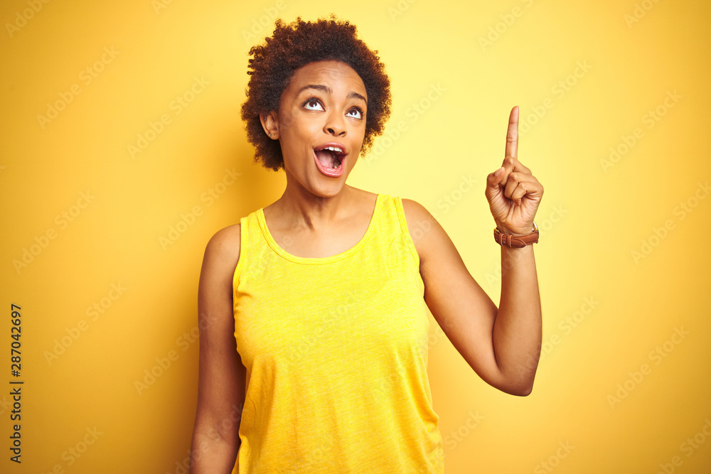 Beauitul african american woman wearing summer t-shirt over isolated yellow background pointing finger up with successful idea. Exited and happy. Number one. Beauitul african american woman wearing summer t-shirt over isolated yellow background pointing finger up with successful idea. Exited and happy. Number one.