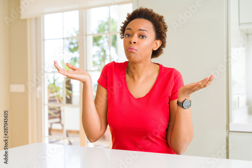 Young beautiful african american woman at home clueless and confused expression with arms and hands raised. Doubt concept.