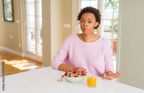 Young african american woman having healthy breakfast in the morning at home making fish face with lips  crazy and comical gesture. Funny expression.