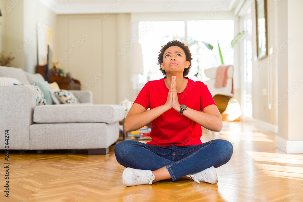 Young beautiful african american woman sitting on the floor at home begging and praying with hands together with hope expression on face very emotional and worried. Asking for forgiveness. Young beautiful african american woman sitting on the floor at home begging and praying with hands together with hope expression on face very emotional and worried. Asking for forgiveness.