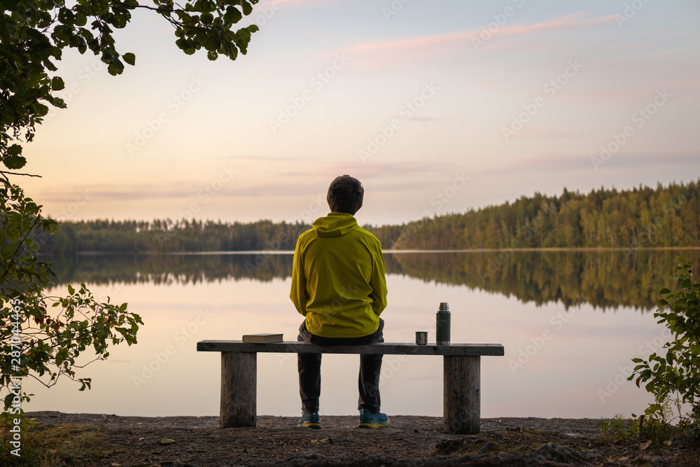 A man in a yellow jacket sits on a bench by the lake on a calm autumn evening. Near a thermos and a book. Blurred background A man in a yellow jacket sits on a bench by the lake on a calm autumn evening. Near a thermos and a book. Blurred background