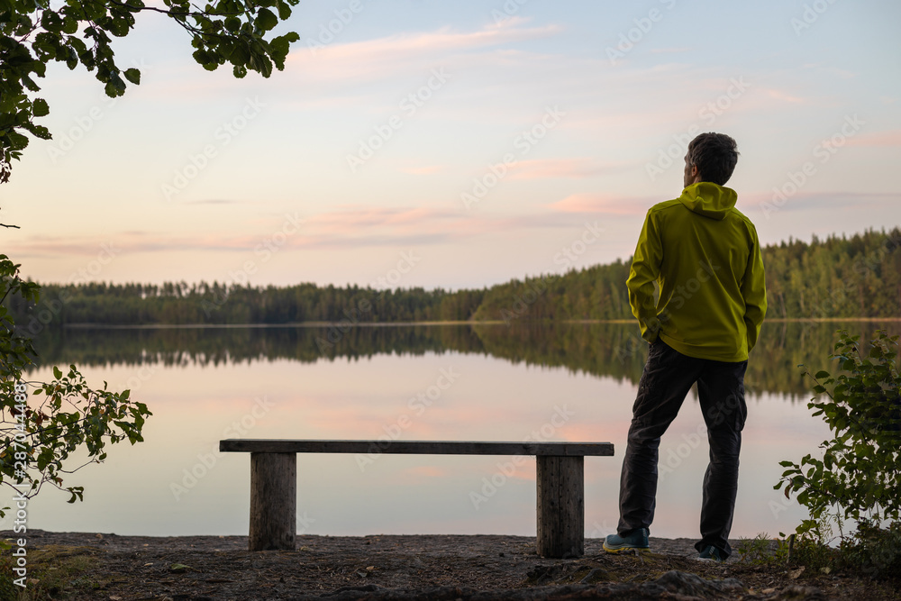 A man in a yellow jacket is standing by a bench on the lake on an autumn evening under the trees. Blurred background