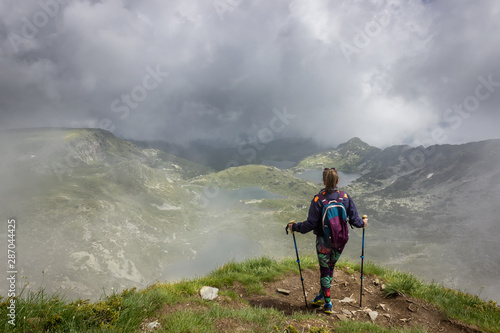 A girl with colorful tights, backpack and walking sticks standing on the edge of Lake peak on Rila mountain, famous viewpoint of seven Rila lakes and looking at iconic lakes through the mist