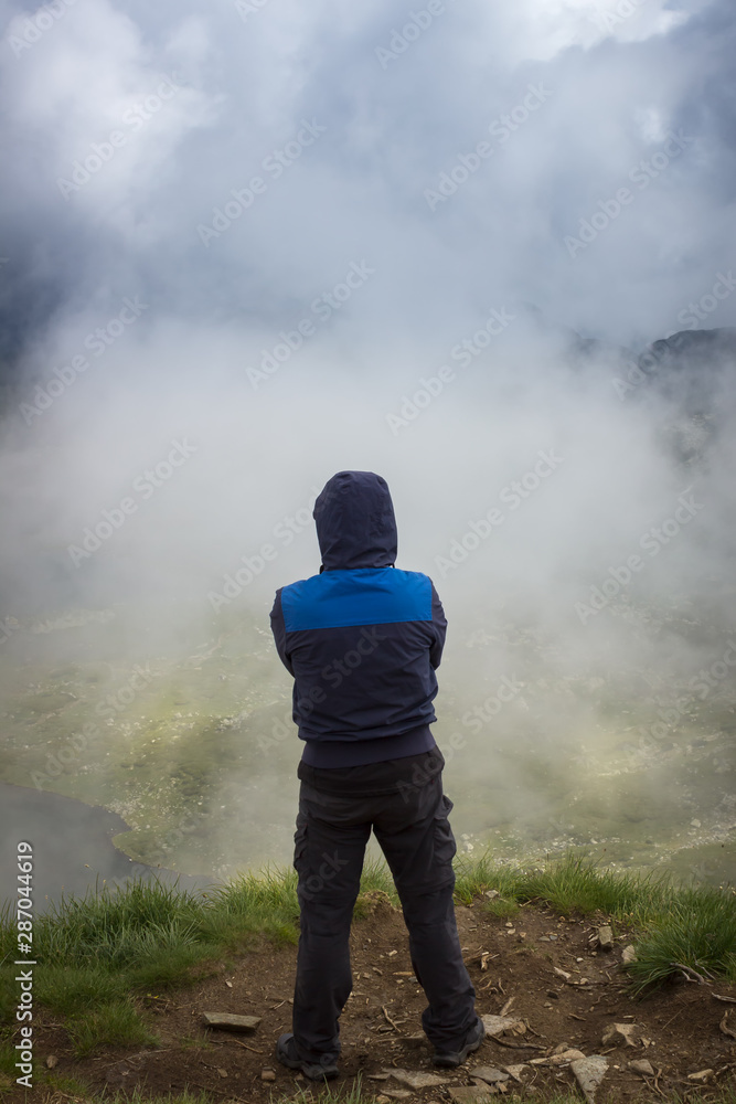 Obraz premium Hiker with blue jacket and hood over head standing at the edge of Lake peak and looking at seven Rila lakes through thick fog