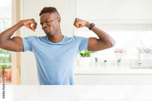 Handsome african american man wearing casual t-shirt at home showing arms muscles smiling proud. Fitness concept.