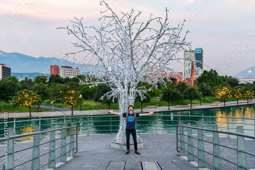 A man standing and spreading his arms in front of a silver lights tree in Monterrey, Mexico