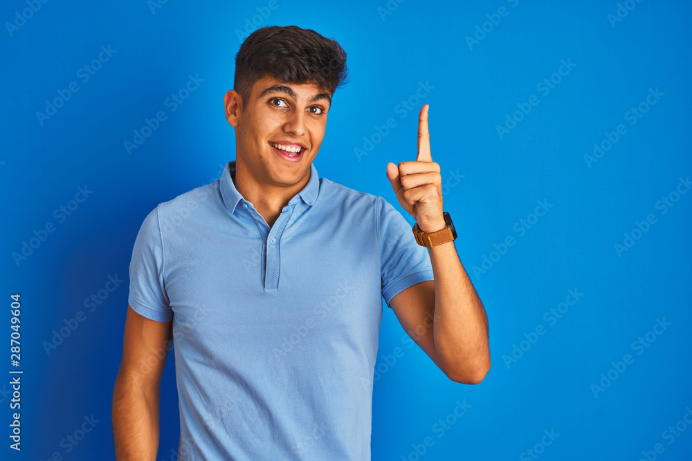 Fototapeta premium Young indian man wearing casual polo standing over isolated blue background pointing finger up with successful idea. Exited and happy. Number one.