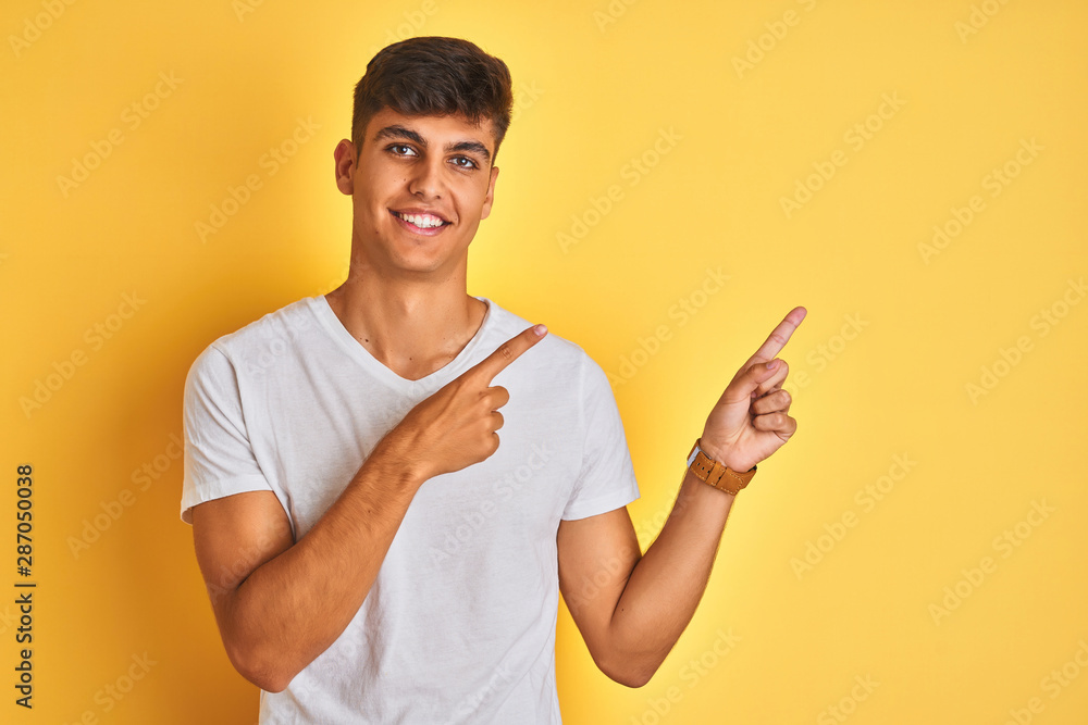 Young indian man wearing white t-shirt standing over isolated yellow background smiling and looking at the camera pointing with two hands and fingers to the side. Young indian man wearing white t-shirt standing over isolated yellow background smiling and looking at the camera pointing with two hands and fingers to the side.