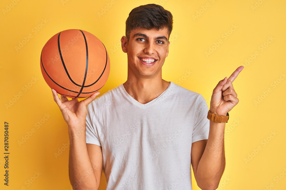 Young indian sportsman holding basketball ball standing over isolated yellow background very happy pointing with hand and finger to the side