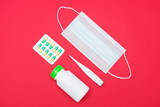 Red desk, medical mask, pills in blister pack, bottle with pills, thermometer, top view, red background