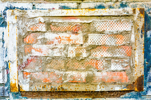 On old brick wall is insert of other bricks in rusty metal frame, peeling paint