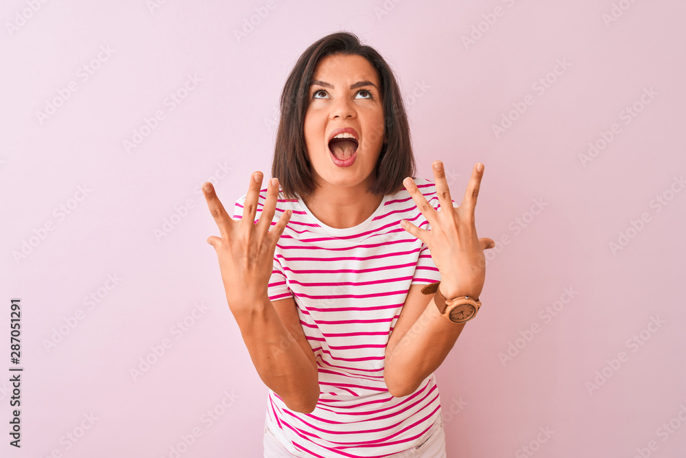 Young beautiful woman wearing striped t-shirt standing over isolated pink background crazy and mad shouting and yelling with aggressive expression and arms raised. Frustration concept. Young beautiful woman wearing striped t-shirt standing over isolated pink background crazy and mad shouting and yelling with aggressive expression and arms raised. Frustration concept.
