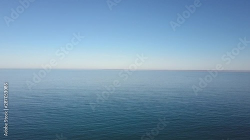 Aerial view of drone flight rising above calm and tranquil ocean water, with summer sunny blue sky and horizon as background and copy space.
