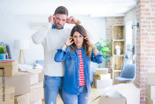 Young beautiful couple standing at new home around cardboard boxes covering ears with fingers with annoyed expression for the noise of loud music. Deaf concept.