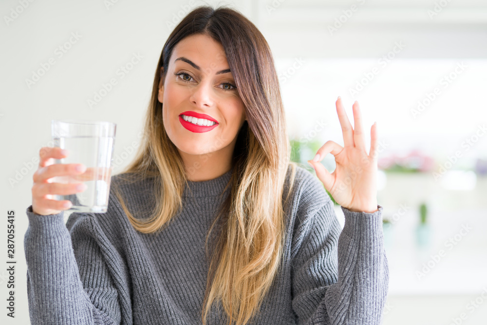 Young beautiful woman drinking glass of water at home doing ok sign with fingers, excellent symbol Young beautiful woman drinking glass of water at home doing ok sign with fingers, excellent symbol