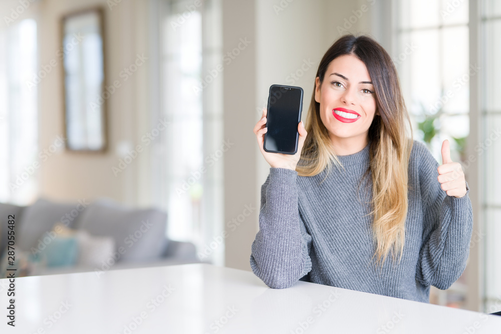 Young beautiful woman showing smartphone screen at home happy with big smile doing ok sign, thumb up with fingers, excellent sign Young beautiful woman showing smartphone screen at home happy with big smile doing ok sign, thumb up with fingers, excellent sign
