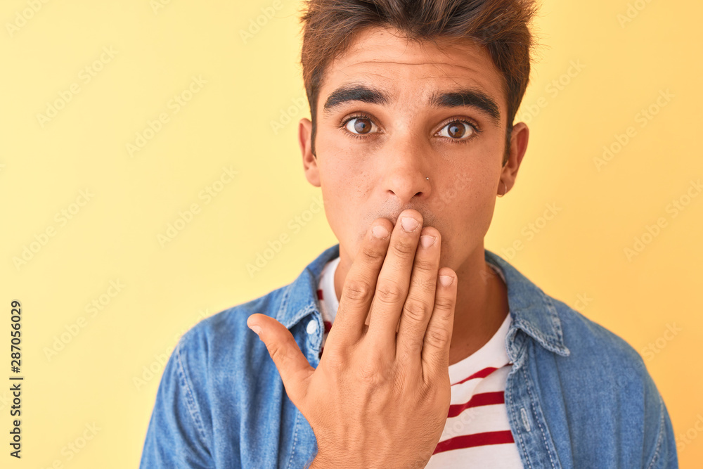 Young handsome man wearing striped t-shirt and denim shirt over isolated yellow background cover mouth with hand shocked with shame for mistake, expression of fear, scared in silence, secret concept Young handsome man wearing striped t-shirt and denim shirt over isolated yellow background cover mouth with hand shocked with shame for mistake, expression of fear, scared in silence, secret concept