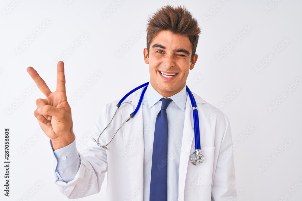 Young handsome doctor man wearing stethoscope over isolated white background smiling with happy face winking at the camera doing victory sign. Number two. Young handsome doctor man wearing stethoscope over isolated white background smiling with happy face winking at the camera doing victory sign. Number two.