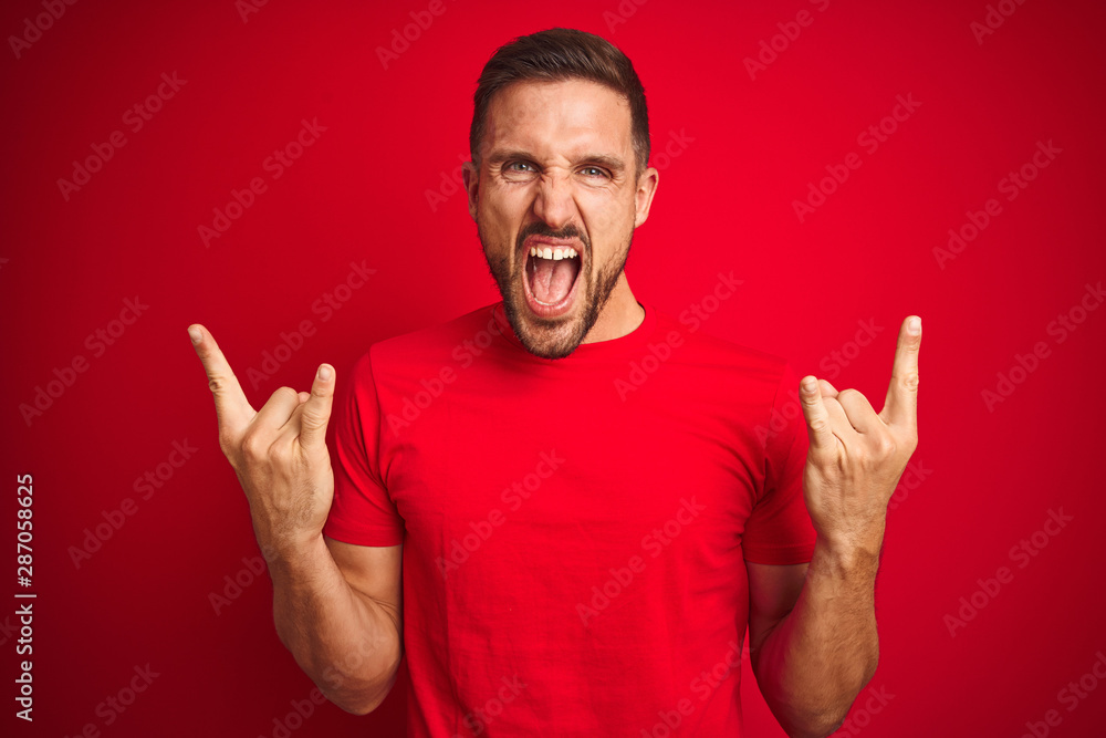 Young handsome man wearing casual t-shirt over red isolated background shouting with crazy expression doing rock symbol with hands up. Music star. Heavy music concept. Young handsome man wearing casual t-shirt over red isolated background shouting with crazy expression doing rock symbol with hands up. Music star. Heavy music concept.