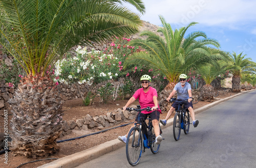 A senior couple man and woman enjoying freedom with electric bicycle. Happiness, fun and emotion outdoor in the street. Flowering plants and palm trees in background