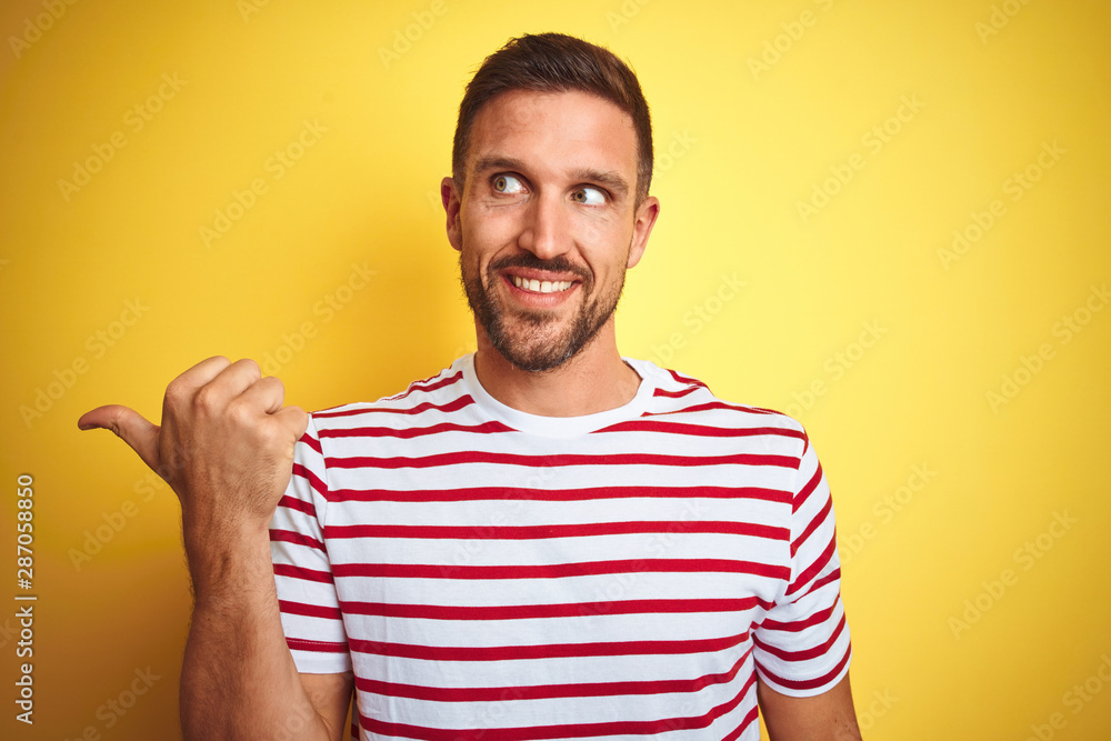 Young handsome man wearing casual red striped t-shirt over yellow isolated background smiling with happy face looking and pointing to the side with thumb up. Young handsome man wearing casual red striped t-shirt over yellow isolated background smiling with happy face looking and pointing to the side with thumb up.