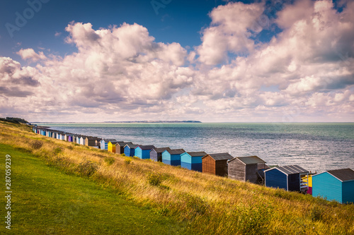 Row of beach huts under a white cloudscape along the coast in Tankerton, Whitstable, Kent. The grass slopes behind the huts are turning a golden yellow. Sheppey can be seen along the horizon. photo