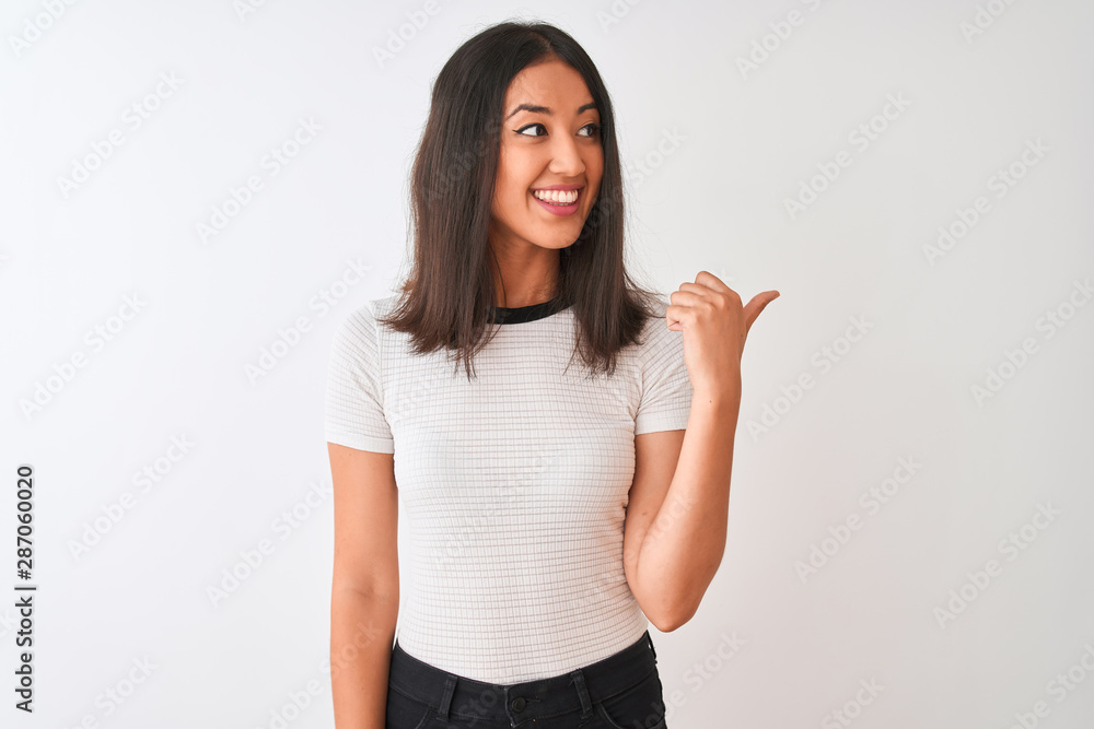 Young beautiful chinese woman wearing casual t-shirt standing over isolated white background smiling with happy face looking and pointing to the side with thumb up. Young beautiful chinese woman wearing casual t-shirt standing over isolated white background smiling with happy face looking and pointing to the side with thumb up.