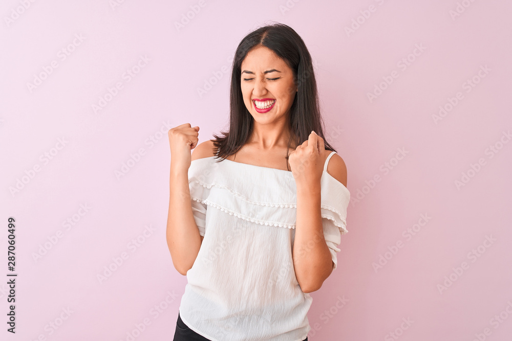 Beautiful chinese woman wearing white t-shirt standing over isolated pink background very happy and excited doing winner gesture with arms raised, smiling and screaming for success. Beautiful chinese woman wearing white t-shirt standing over isolated pink background very happy and excited doing winner gesture with arms raised, smiling and screaming for success.
