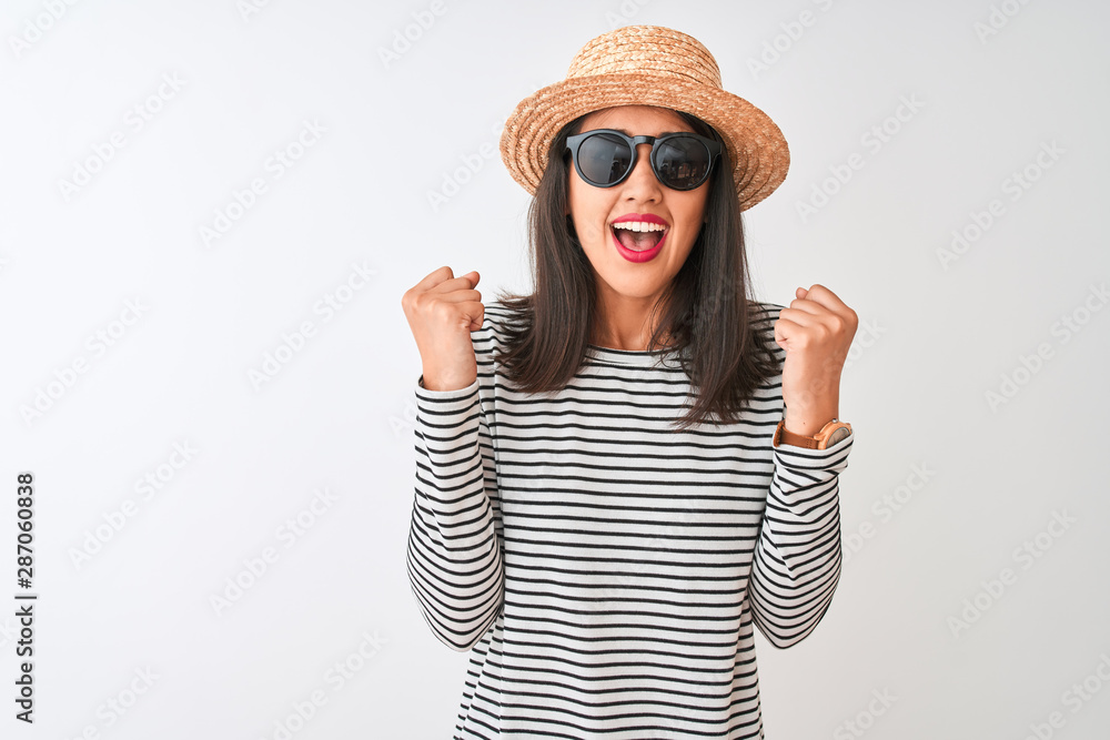 Chinese woman wearing striped t-shirt hat sunglasses standing over isolated white background celebrating surprised and amazed for success with arms raised and open eyes. Winner concept. Chinese woman wearing striped t-shirt hat sunglasses standing over isolated white background celebrating surprised and amazed for success with arms raised and open eyes. Winner concept.