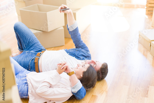Beautiful couple hugging in love on the floor, relaxing and smiling taking a selfie with smartphone at new apartment around cardboard boxes