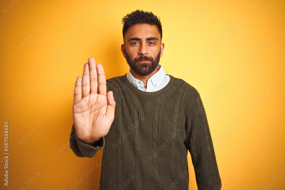 Young indian man wearing green sweater and shirt standing over isolated yellow background doing stop sing with palm of the hand. Warning expression with negative and serious gesture on the face. Young indian man wearing green sweater and shirt standing over isolated yellow background doing stop sing with palm of the hand. Warning expression with negative and serious gesture on the face.