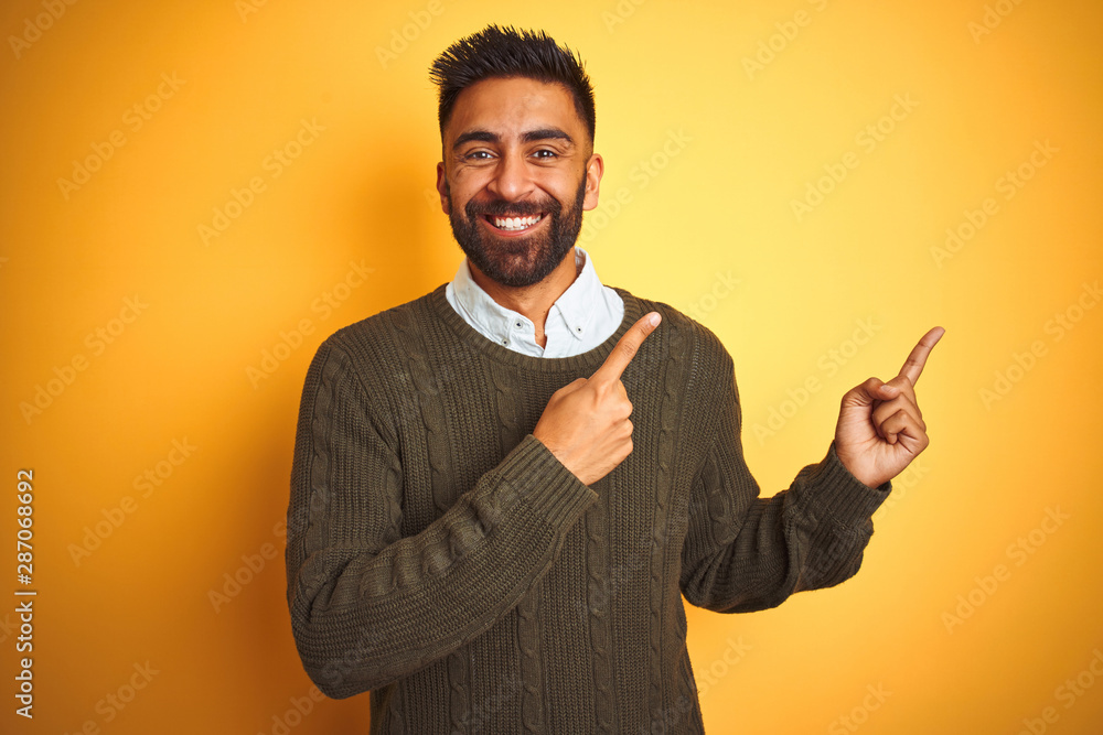 Young indian man wearing green sweater and shirt standing over isolated yellow background smiling and looking at the camera pointing with two hands and fingers to the side. Young indian man wearing green sweater and shirt standing over isolated yellow background smiling and looking at the camera pointing with two hands and fingers to the side.
