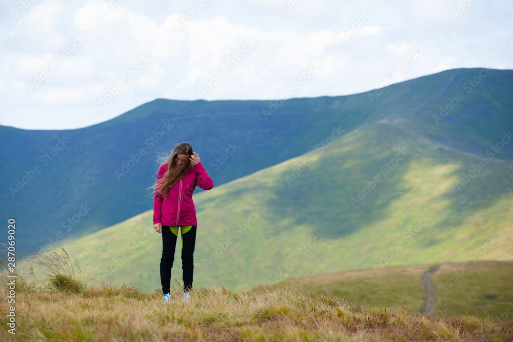 Happy young girl hiker in a red sweater standing on the background of mountain while her hair fluttering in the wind