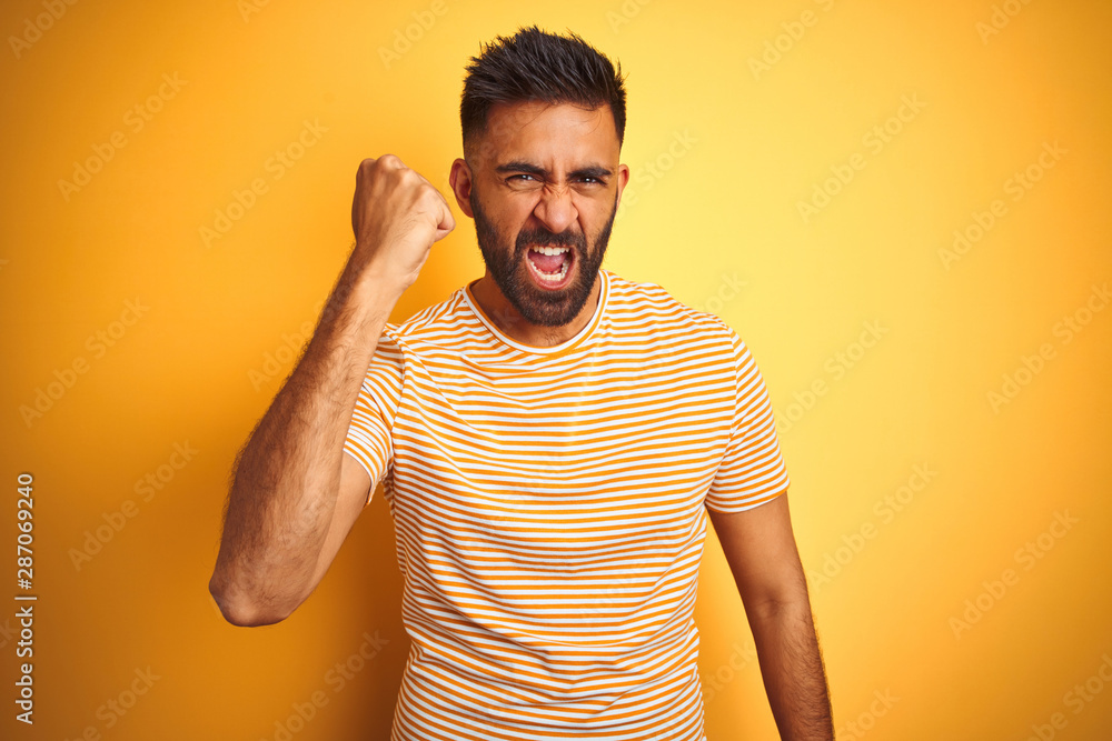 Young indian man wearing t-shirt standing over isolated yellow background angry and mad raising fist frustrated and furious while shouting with anger. Rage and aggressive concept. Young indian man wearing t-shirt standing over isolated yellow background angry and mad raising fist frustrated and furious while shouting with anger. Rage and aggressive concept.