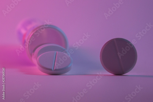 White round pill near spilled from the jar in the background with a pink light.One pill stands on the rib next to it. The concept of treatment, medications, disease