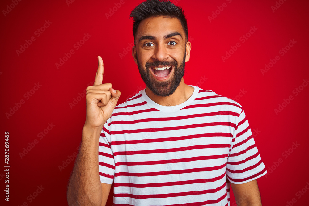Fototapeta premium Young indian man wearing striped t-shirt standing over isolated red background pointing finger up with successful idea. Exited and happy. Number one.