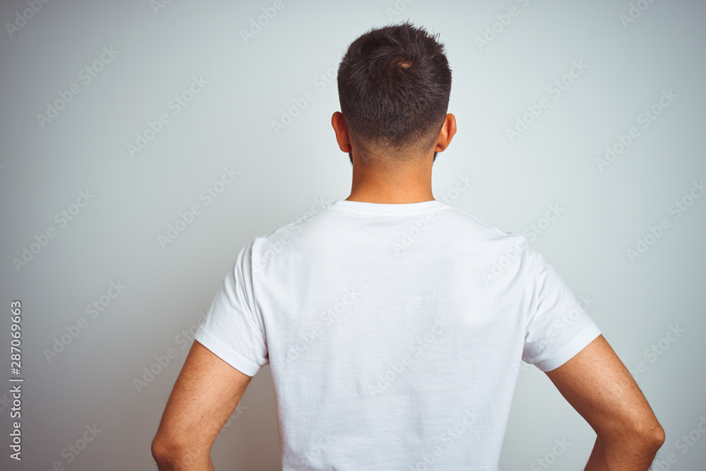 Young indian man wearing t-shirt standing over isolated white background standing backwards looking away with arms on body - obrazy, fototapety, plakaty 