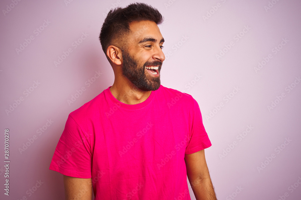 Young indian man wearing t-shirt standing over isolated pink background looking away to side with smile on face, natural expression. Laughing confident. Young indian man wearing t-shirt standing over isolated pink background looking away to side with smile on face, natural expression. Laughing confident.