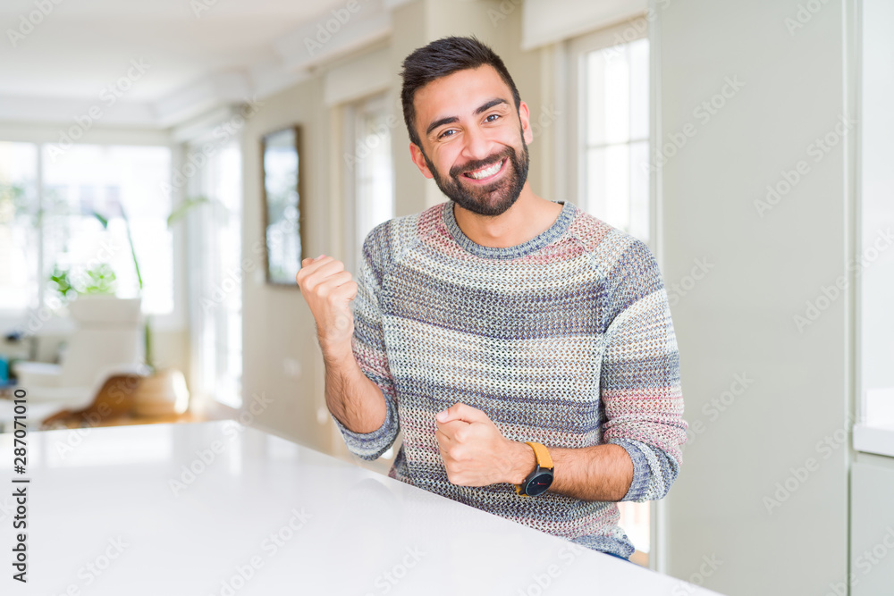 Fototapeta premium Handsome hispanic man wearing casual sweater at home very happy and excited doing winner gesture with arms raised, smiling and screaming for success. Celebration concept.