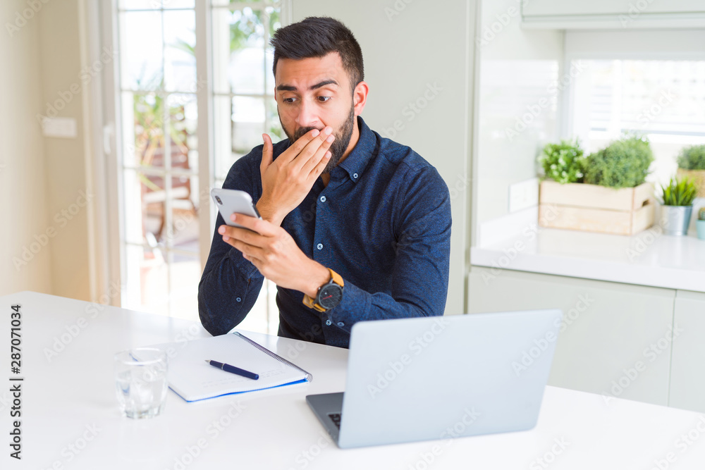 Handsome hispanic business man using smartphone and laptop at the office cover mouth with hand shocked with shame for mistake, expression of fear, scared in silence, secret concept Handsome hispanic business man using smartphone and laptop at the office cover mouth with hand shocked with shame for mistake, expression of fear, scared in silence, secret concept