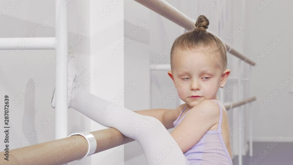 Adorable little ballerina girl putting her leg on ballet bar for stretching. Happy little girl in leotard exercising at ballet school, stretching her leg on barre. Flexibility, gymnastics concept Adorable little ballerina girl putting her leg on ballet bar for stretching. Happy little girl in leotard exercising at ballet school, stretching her leg on barre. Flexibility, gymnastics concept